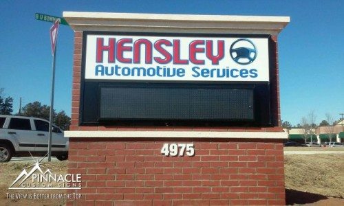 Monument sign for Hensley Automotive Services, created by Pinnacle Custom Signs. The sign displays the company name and logo in bold red and blue letters, mounted on a brick base with a digital display panel below. Positioned at 4975, the sign enhances roadside visibility and business branding.