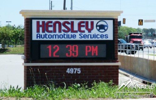 Digital monument sign for Hensley Automotive Services, designed by Pinnacle Custom Signs. The sign features the company name and logo in bold letters above a digital display showing the time. Mounted on a brick base, the sign is positioned by the roadside, providing clear visibility to passing traffic and enhancing business recognition.