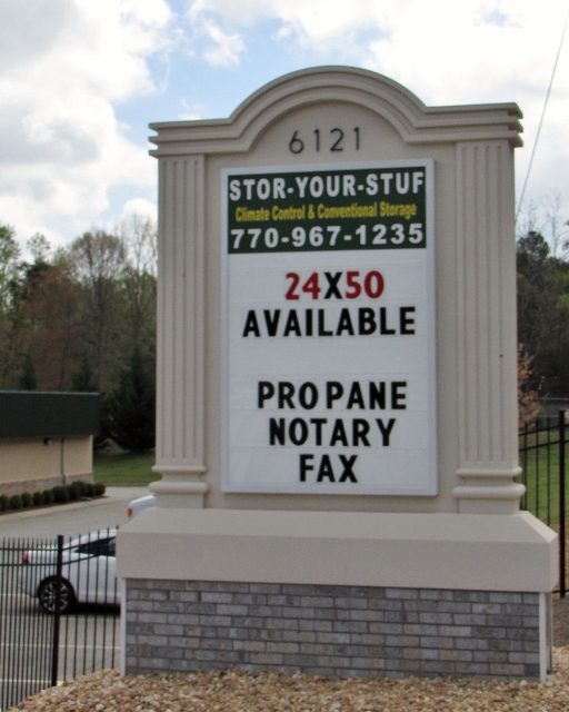 Monument sign for Stor-Your-Stuf Climate Control & Conventional Storage, created by Pinnacle Custom Signs. The sign displays the business name, phone number, and additional services like propane, notary, and fax. It features a classic design with white pillars and a brick base, situated at 6121, enhancing roadside visibility and providing essential information.