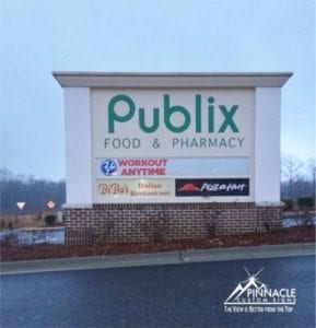 A large monument sign for 'Publix Food & Pharmacy' with additional business signs for 'Workout Anytime,' 'Dr. Bob’s Italian Restaurant,' and 'Pizza Hut' displayed below it. The sign is set against a landscaped background with shrubs and a brick base. The Pinnacle Custom Signs logo and tagline 'The View is Better from the Top' are in the bottom right corner of the image.