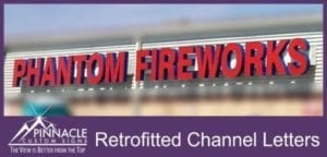 Red channel letters spelling 'PHANTOM FIREWORKS' mounted on the exterior of a building. The letters are large, bold, and prominently displayed. The image is labeled 'Retrofitted Channel Letters' with the Pinnacle Custom Signs logo and the tagline 'The View is Better from the Top' at the bottom. The sign is designed to attract attention and is likely illuminated for visibility.