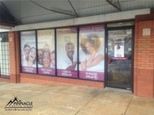 Colorful window graphics for a storefront, designed by Pinnacle Custom Signs, featuring images of diverse, smiling individuals along with text promoting dental services. The graphics cover several windows, enhancing the visibility and appeal of the dental practice.