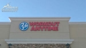 Exterior view of a business sign for '24/7 Workout Anytime' mounted on a beige building with a stone accent wall. The sign features red capital letters and a circular logo with '24/7' in blue and white. The Pinnacle Custom Signs logo and tagline 'The View is Better from the Top' are in the top left corner of the image.