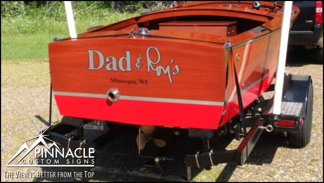 A boat with a custom wrap that reads 'Dad & Ron's' with 'Minocqua, WI' below it, applied to the stern. The wrap features elegant, easy-to-read white lettering on a wooden background. The boat is on a trailer, showcasing the professional design and application work by Pinnacle Custom Signs.