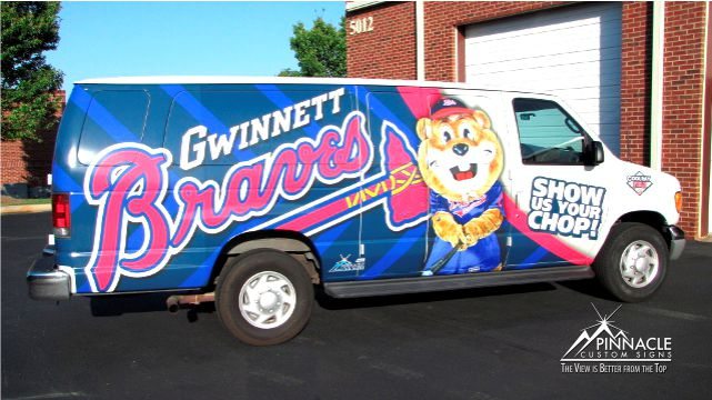 Van with custom wrap by Pinnacle Custom Signs, advertising the Gwinnett Braves, featuring the team logo, mascot, and the slogan 'Show Us Your Chop!' parked near a brick building