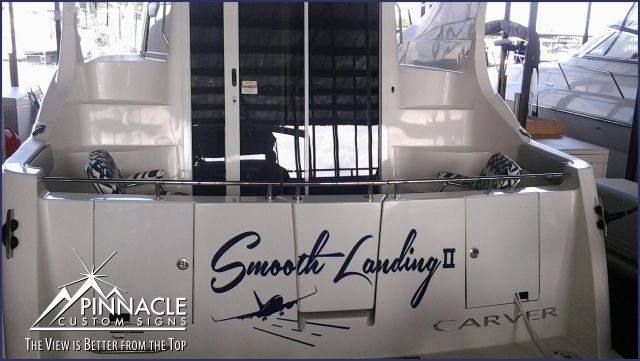 A close-up view of a boat's stern with a custom wrap displaying the name 'Smooth Landing II' in blue, stylized letters. Below the name is an image of a plane in flight. The boat is docked in a marina, and the wrap is applied neatly, showcasing the professional quality of Pinnacle Custom Signs.