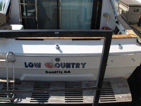 A boat at a dock with a custom wrap text on the stern reading 'Low Country' with a small crab graphic integrated into the text. Below it, the location 'Sandfly, GA' is displayed in bold black letters. The boat is positioned in a covered dock area with a wooden platform, showcasing the professional boat wrap by Pinnacle Custom Signs.