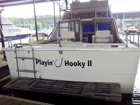 A boat docked at a marina, showing the stern with custom wrap text 'Playin' Hooky II' in bold black letters. The boat features a fishing hook integrated into the text design. The boat is positioned in a covered dock area, surrounded by other boats and watercraft, demonstrating the professional boat wrap by Pinnacle Custom Signs.