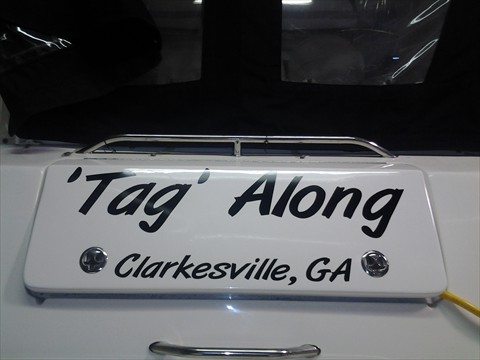A close-up of a boat with a custom wrap displaying the name 'Tag Along' in black, stylized letters on a white background. Below, it reads 'Clarkesville, GA.' The wrap is applied neatly on the stern of the boat, demonstrating the clean and professional work by Pinnacle Custom Signs.