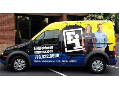 Van with custom wrap by Pinnacle Custom Signs, advertising Embroidered Impressions, featuring the company logo, contact information, and images of two people in branded clothing, parked near a brick building