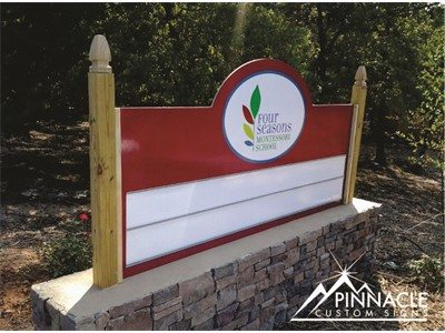 An outdoor location sign for Four Seasons Montessori School, featuring a red background with white spaces for additional information. The sign is supported by two wooden posts and sits on a stone base. The logo of Four Seasons Montessori School is prominently displayed at the top center of the sign. The image is branded with the Pinnacle Custom Signs logo in the bottom right corner.