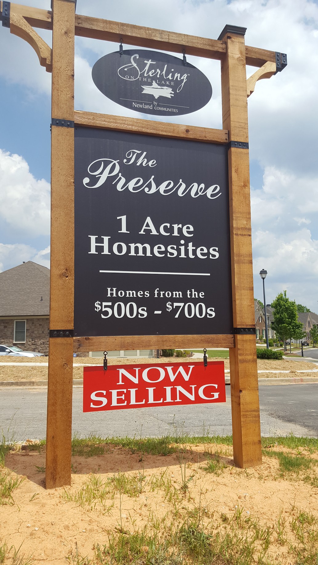 Custom Post & Panel Signs | Post Signs For Real Estate Outdoor wooden sign advertising 'Sterling on the Lake by Newland Communities' with 'The Preserve' in elegant script. The sign mentions '1 Acre Homesites' with homes priced from '$500s - $700s.' Below, a red banner states 'NOW SELLING.' The sign is set in a residential area with homes and a clear sky in the background.