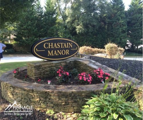 Stone Monument Sign | Monument Sign Design Monument sign for 'Chastain Manor,' featuring a black oval sign with gold lettering. The sign is mounted on a low stone wall with a raised planter bed, containing pink flowers and greenery. The surrounding area includes a well-maintained lawn and tall evergreen trees. The sign is created by Pinnacle Custom Signs.