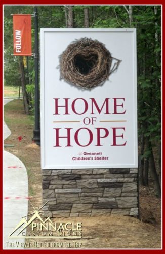 Monument Signs Cost | Stone Monument Signs Monument sign for 'Home of Hope' at Gwinnett Children's Shelter, featuring a large nest graphic at the top. The sign is mounted on a stone base and surrounded by greenery, with a visible pathway and a 'FOLLOW' sign in the background. The sign is created by Pinnacle Custom Signs.