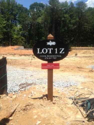 Residential Lot Signs | Commercial Lot Signs Wooden sign marking 'Lot 1Z' by Cook Residential with a contact number. Below, a red banner invites to 'Visit the Information Center.' The sign is placed on a construction site with cleared land, gravel, and some construction materials visible. The background features a forested area with dense trees and a clear sky.