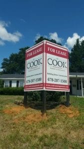 Large commercial property sign with 'For Lease' in red banners at the top and bottom. The central part of the sign displays 'Cook' and 'Building Better Communities' in large text, with a contact number. The sign is mounted on black posts and stands on a grassy area with a building and trees in the background under a clear blue sky.