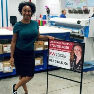 An African-American woman standing next to a yard sign in a workshop. The yard sign features a photo of a woman and text that reads 'Courtney Murphy, KW Atlanta Partners' and a phone number. The background includes various materials and equipment used for sign-making.