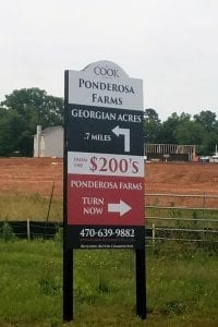 Directional sign for Ponderosa Farms by Cook. The sign indicates 'Georgian Acres 7 miles' with an arrow pointing right. Below it, text reads 'From the $200's' and 'Ponderosa Farms Turn Now' with an arrow pointing right. The contact number is displayed at the bottom. The sign stands in a grassy area with a construction site and trees in the background.