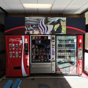 A branded vending machine setup with two Coca-Cola vending machines flanking a central snack vending machine. Above the machines is a large branding sign featuring the logo of Georgia State University along with an image of a panther statue and campus scenes. The overall display promotes the university's branding and partnership with Coca-Cola.
