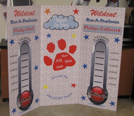 Branding display for a school event called 'Wildcat Run-A-Pawlooza,' featuring a pledge goal and pledges collected thermometers on either side. The center of the display has a large paw print graphic with the words 'Support Our School' and 'Run, Jog, Skip, Walk' around the paw. The thermometers track pledges up to 20,000, with the left labeled 'Goal' and the right labeled 'Collected.'