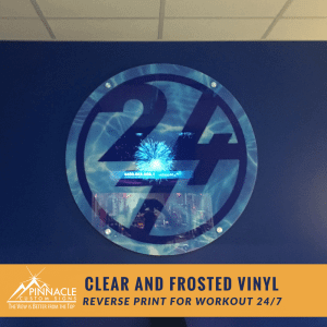 Clear and frosted vinyl sign for 'Workout 24/7,' featuring a reverse print design. The circular sign includes the '24/7' logo and is mounted on a dark blue wall. The sign is created by Pinnacle Custom Signs, with their logo and branding visible at the bottom of the image.