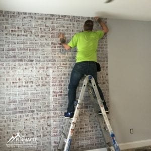 A worker wearing a green shirt stands on a ladder, applying a wall graphic with a brick pattern to a light gray wall. The graphic creates a realistic brick wall effect. The image is branded with the Pinnacle Custom Signs logo in the bottom left corner.
