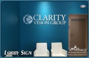 A modern lobby sign for 'Clarity Vision Group' mounted on a blue wall. The sign includes the company logo and name in white. Below the sign, there are two white chairs placed against the wall. The image also contains a watermark of Pinnacle Custom Signs with the tagline 'The View is Better from the Top' in the bottom right corner.