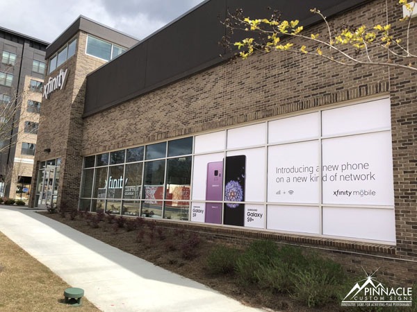 Promotional window graphics for Xfinity Mobile, designed by Pinnacle Custom Signs, featuring large advertisements for the Samsung Galaxy S9 and S9+ on a brick storefront. The graphics cover several windows, providing high visibility and effectively promoting the new phone and network.