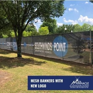 A large mesh banner displayed on a fence, featuring the text 'Southwinds Point, Apartment Homes' with a new logo. The banner is located outdoors, surrounded by trees and greenery. The image includes a blue banner at the bottom right corner with the text 'Mesh Banners with New Logo' and the Pinnacle Custom Signs logo.