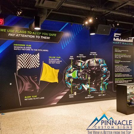 Indoor wall graphics for AMP Kart Racing, featuring information on flag signals and a detailed diagram of a go-kart engine, displayed in a brightly lit exhibition space
