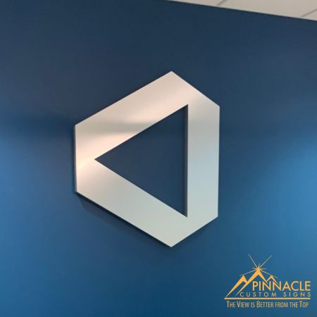 Modern lobby sign featuring a geometric logo, designed by Pinnacle Custom Signs, mounted on a blue wall. The minimalist design of the white, three-dimensional shape creates a sleek and contemporary look, enhancing the professional atmosphere of the office.